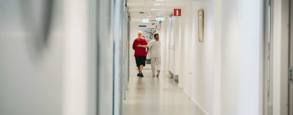 Äldre herre går genom en korridor med en behandlare vid sin sida